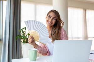 fatigué surchauffé Jeune femme tenir vague ventilateur souffrir de chaleur transpiration intérieur travail sur portable à Accueil bureau, agacé femme ressentir inconfortable chaud été temps problème non air Conditionneur concept photo