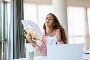 fatigué surchauffé Jeune femme tenir vague ventilateur souffrir de chaleur transpiration intérieur travail sur portable à Accueil bureau, agacé femme ressentir inconfortable chaud été temps problème non air Conditionneur concept photo