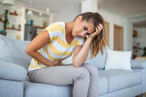 femme allongé sur le canapé, apparaissant indisposé. magnifique Jeune Dame mensonge sur le lit, doucement étreinte sa estomac. femme expérience une douloureux maux d'estomac tandis que sur le lit, pendant sa menstruel période photo