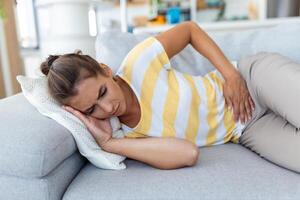 femme allongée sur un canapé qui a l'air malade dans le salon. belle jeune femme allongée sur le lit et se tenant la main sur le ventre. femme ayant des maux d'estomac douloureux au lit, période menstruelle photo