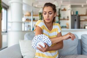 Jeune femme est séance à Accueil sur sa canapé et émouvant le sa coude tandis que Souffrance de coude douleur, placement la glace pack sur blesser place photo