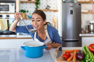 content Jeune femme cuisine dégustation dîner dans une pot permanent dans moderne cuisine à maison. femme au foyer en train de préparer en bonne santé nourriture souriant . Ménage et nutrition. suivre un régime recettes concept photo