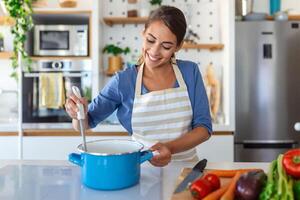 content Jeune femme cuisine dégustation dîner dans une pot permanent dans moderne cuisine à maison. femme au foyer en train de préparer en bonne santé nourriture souriant . Ménage et nutrition. suivre un régime recettes concept photo