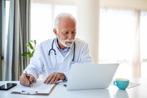 sérieux mature médecin en utilisant portable et séance à bureau. Sénior professionnel médical médecin portant blanc manteau et stéthoscope travail sur ordinateur à lieu de travail. photo