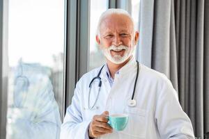 portrait de Sénior mature santé se soucier professionnel, médecin, avec stéthoscope en portant bleu tasse de café, photo