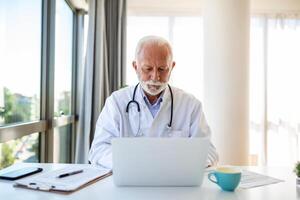 sérieux mature médecin en utilisant portable et séance à bureau. Sénior professionnel médical médecin portant blanc manteau et stéthoscope travail sur ordinateur à lieu de travail. photo