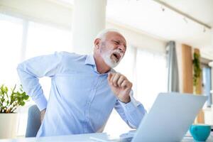 Sénior homme d'affaire travail séance à bureau souffre de inférieur retour douleur. dommage de intervertébral disques, spinal les articulations, compression de nerf les racines causé par faux posture et sédentaire travail photo