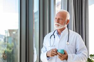 portrait de Sénior mature santé se soucier professionnel, médecin, avec stéthoscope en portant bleu tasse de café, photo