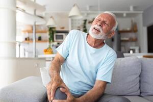 le plus âgée homme est séance sur le canapé à maison, a douleur dans le le genou articulation, en portant le sien jambe, arthrose concept. photo