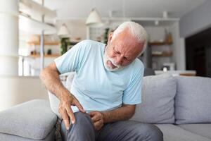 le plus âgée homme est séance sur le canapé à maison, a douleur dans le le genou articulation, en portant le sien jambe, arthrose concept. photo