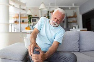 le plus âgée homme est séance sur le canapé à maison, a douleur dans le le genou articulation, en portant le sien jambe, arthrose concept. photo