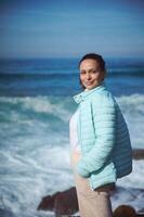 souriant femme permanent par océan sur le rocheux falaise, à la recherche à caméra, profiter le vue de magnifique vagues photo