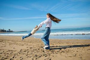 plein longueur portrait de une content mère filage sa peu enfant fille, un adorable fille tandis que en jouant sur le plage photo