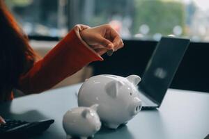 une Jeune fille travaux à une ordinateur portable, écrit une affaires développement plan dans une carnet de notes, développe une stratégie et fait du calculs de gagné fonds. une femme met sa des économies dans une porcin banque. photo