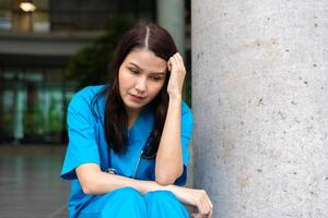 portrait de stressé et surmené femelle médecin médical ouvrier chirurgien séance sur le sol près le en fonctionnement pièce dans le hôpital et prendre de le masque photo