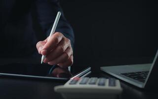 homme d'affaire dans noir costume travail sur portable ordinateur et tablette et calculatrice, main mains toucher sur tablette à Bureau avec foncé arrière-plan, en ligne fonctionnement, proche en haut, copie espace. photo