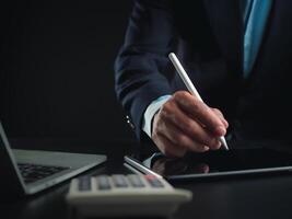 homme d'affaire dans noir costume travail sur portable ordinateur et tablette et calculatrice, main mains toucher sur tablette à Bureau avec foncé arrière-plan, en ligne fonctionnement, proche en haut, copie espace. photo