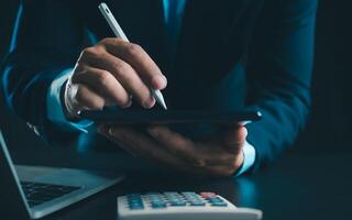 homme d'affaire dans noir costume travail sur portable ordinateur et tablette et calculatrice, main mains toucher sur tablette à Bureau avec foncé arrière-plan, en ligne fonctionnement, proche en haut, copie espace. photo