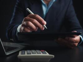 homme d'affaire dans noir costume travail sur portable ordinateur et tablette et calculatrice, main mains toucher sur tablette à Bureau avec foncé arrière-plan, en ligne fonctionnement, proche en haut, copie espace. photo