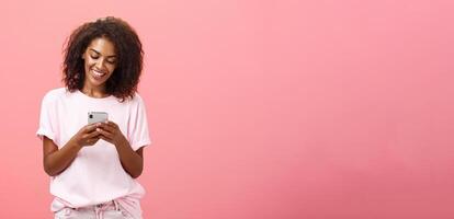 fille l'écriture texte message à ami. portrait de charmant amusé et content Jeune peau foncée femme avec frisé cheveux à la recherche à téléphone intelligent écran dactylographie souriant profiter cool Caractéristiques de téléphone portable photo