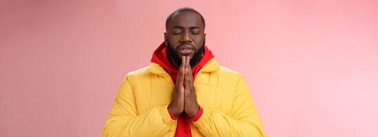 l'air sérieux fidèle Jeune déterminé Afro-américain prier gars dans Jaune manteau presse paumes ensemble prier proche yeux chuchotement supplication demander Dieu Aidez-moi faire souhait, permanent rose Contexte photo