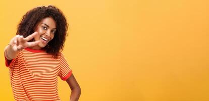 Hey paix mon ami. charmant sortant et sur de soi insouciant peau foncée fille avec afro coiffure dans branché T-shirt tirant main avec la victoire geste vers caméra souriant plus de Orange mur photo