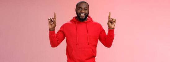 sournois mignonne Afro-américain barbu gars dans rouge sweat à capuche souriant mystérieux connaître exactement quoi vous vouloir montrer du doigt élevé indice les doigts en haut sourire spectacle parfait copie espace promo, rose Contexte photo