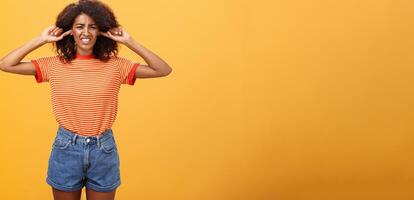 quoi cette ennuyeux son. perturbé et mécontent peau foncée fille avec frisé cheveux renfrogné serrant les dents de malaise fermeture oreilles avec indice les doigts de bruyant irritant du son plus de Orange mur photo