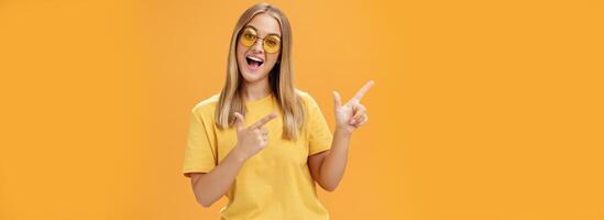 Cliquez sur sa regarder mon vidéos. content sur de soi et sortant charmant Jeune fille avec tout droit cheveux dans des lunettes de soleil et Jaune T-shirt montrer du doigt à plus haut la gauche coin promouvoir produit avec joyeux sourire photo