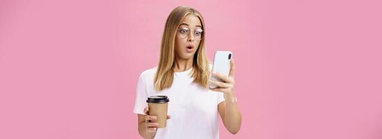 femme en buvant café étant sous le choc par reçu message réagir sur étourdissant nouvelles pliant lèvres halètement à la recherche étonné et impressionné à téléphone intelligent écran en portant papier tasse, posant plus de rose mur photo