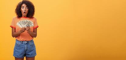 femme sous le choc découverte beaucoup de en espèces dans sûr. portrait de surpris muet beau peau foncée femelle avec frisé la Coupe de cheveux pliant lèvres halètement en portant argent posant plus de Orange Contexte photo