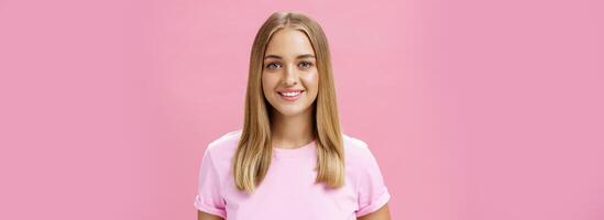 fermer coup de amical agréable commun caucasien fille avec juste cheveux et bronzé peau dans T-shirt souriant largement avec bien blanc les dents regarder à caméra ambitieux et Enchanté plus de rose Contexte photo