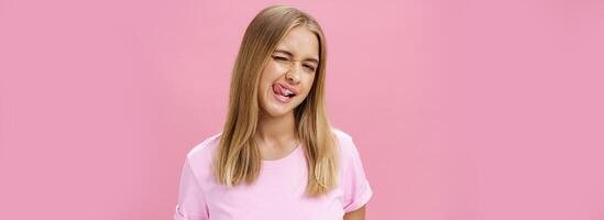 espiègle affectueux attrayant Jeune fille avec juste tout droit cheveux dans T-shirt un clin d'oeil à caméra souriant et lécher plus haut lèvre avec langue sentiment enthousiaste et insouciant contre rose Contexte photo