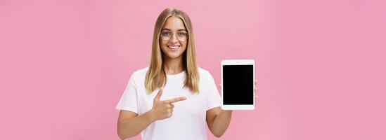 à la taille coup de optimiste et joyeux femelle montrant cool numérique tablette montrer du doigt à dispositif écran et souriant largement à caméra donnant Conseil quoi app utile posant dans des lunettes contre rose mur photo