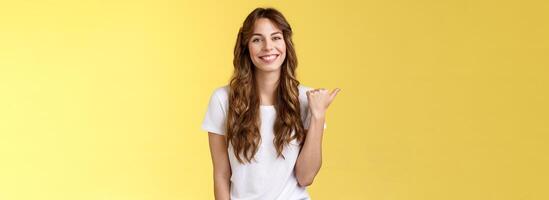 visite voir toi-même. de bonne humeur charismatique beau sortant fille longue frisé la Coupe de cheveux montrant endroit faire bien coiffure souriant Heureusement Enchanté montrer du doigt pouce la gauche présenter promo Jaune Contexte photo