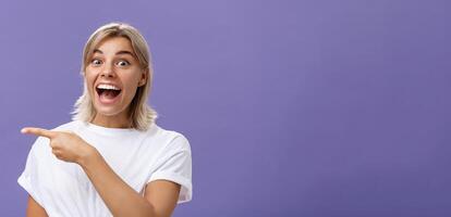 à la taille coup de joyeux ravi attrayant et sensible Jeune blond femelle avec bronzé peau dans blanc décontractée T-shirt souriant et en riant Heureusement montrer du doigt la gauche avec excitation plus de violet mur photo