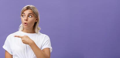 fermer coup de intrigué intéressé attrayant adulte blond femelle dans blanc T-shirt pliant lèvres regarder et montrer du doigt la gauche avec curieuse et ravi expression plus de violet Contexte photo