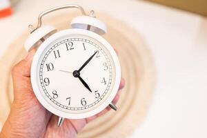 main en portant une blanc alarme l'horloge sur une blanc table et marron Contexte. photo