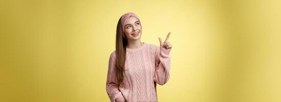 portrait de amusé intéressé mignonne Années 20 européen fille portant chandail, Bandeau à la recherche plus haut la gauche coin montrer du doigt de côté souriant intrigué, en extase, capturé par curieuse promo plus de Jaune mur photo