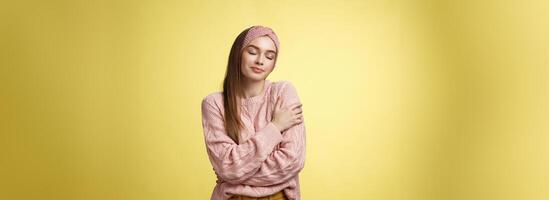 sucré charme Jeune fille portant chaud confortable chandail embrassement se traversée bras plus de corps dans câlin, souriant doux et gentil proche yeux, rappel charmant jours, soumissionner souvenirs plus de Jaune mur photo