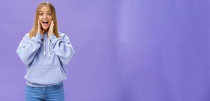 femme joyeusement souriant à caméra et émouvant des joues comme si étant surpris et content voir ami réunion la personne dans aéroport sentiment de bonne humeur à réunir permanent plus de violet Contexte dans mignonne sweat à capuche photo