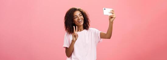 élégant sociable beau peau foncée femelle étudiant avec frisé coiffure tirant main avec téléphone intelligent près visage prise selfie montrant paix signe à dispositif écran tandis que souriant insouciant photo