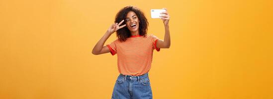 fille fabrication vidéo vlog avec marque Nouveau téléphone intelligent affectation dans l'Internet en essayant devenir célèbre permanent plus de Orange Contexte posant pour selfie regarder à gadget écran montrant paix ou la victoire geste photo