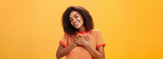 à la taille coup de charmant sensuel peau foncée femme avec frisé coiffure dans branché T-shirt en portant paumes sur cœur à la recherche avec heureux aimant sourire sentiment reconnaissant et Enchanté plus de Orange mur photo