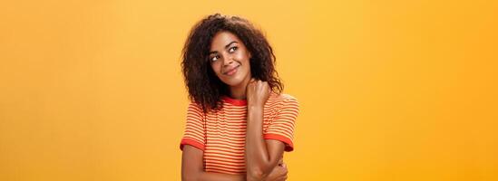 mignonne et doux rêveur peau foncée fille avec cruellement coiffure basculant tête souriant de délice et bonheur émouvant cou soumissionner regarder à plus haut la gauche coin en pensant ou étant nostalgique photo