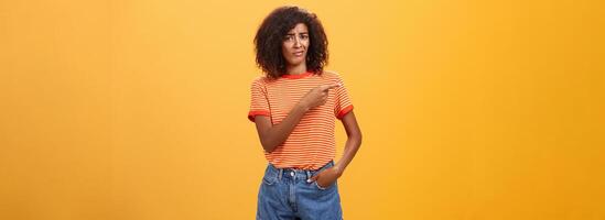 ne pas vaut mon temps. mécontent pas impressionné élégant attrayant peau foncée femelle modèle dans rayé T-shirt et denim short renfrogné avec pointilleux expression montrer du doigt la gauche douteux et mécontent photo