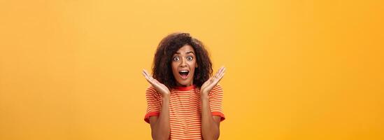 à la taille coup de étonné joyeux sociable peau foncée femelle avec afro coiffure dans rayé T-shirt faire des gestes avec paumes élevé près visage regarder étonné et Enchanté à caméra de surprise et joie photo