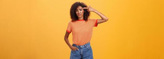 portrait de mécontent sombre et triste agacé peau foncée femelle avec frisé coiffure montrant pistolet avec main près temple renfrogné vouloir à tuer se de échappement et ennui plus de Orange mur photo