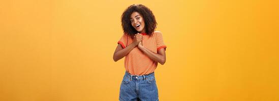 fille soupirs et regarder à mignonne chose serrant paumes ensemble près cœur souriant avec admiration et l'amour étant féminin et soumissionner posant plus de Orange Contexte sentiment affection photo