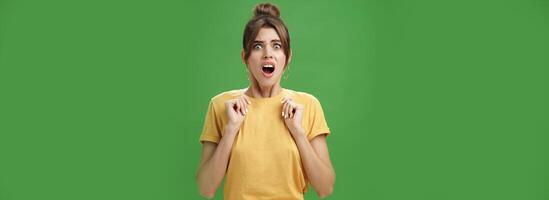 sous le choc féminin charme femme avec peigné cheveux dans Jaune T-shirt renfrogné halètement avec ouvert bouche en portant paumes pressé à poitrine de aversion et dégoûter à la recherche à terrifiant chose plus de vert mur photo